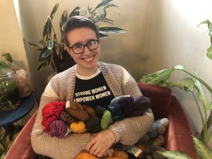 Image Description: A white, queer woman with black framed glasses sits in a red chair, smiling and holding a bunch of brightly colored skeins of yarn. She is surrounded by various sizes of green and white house plants.